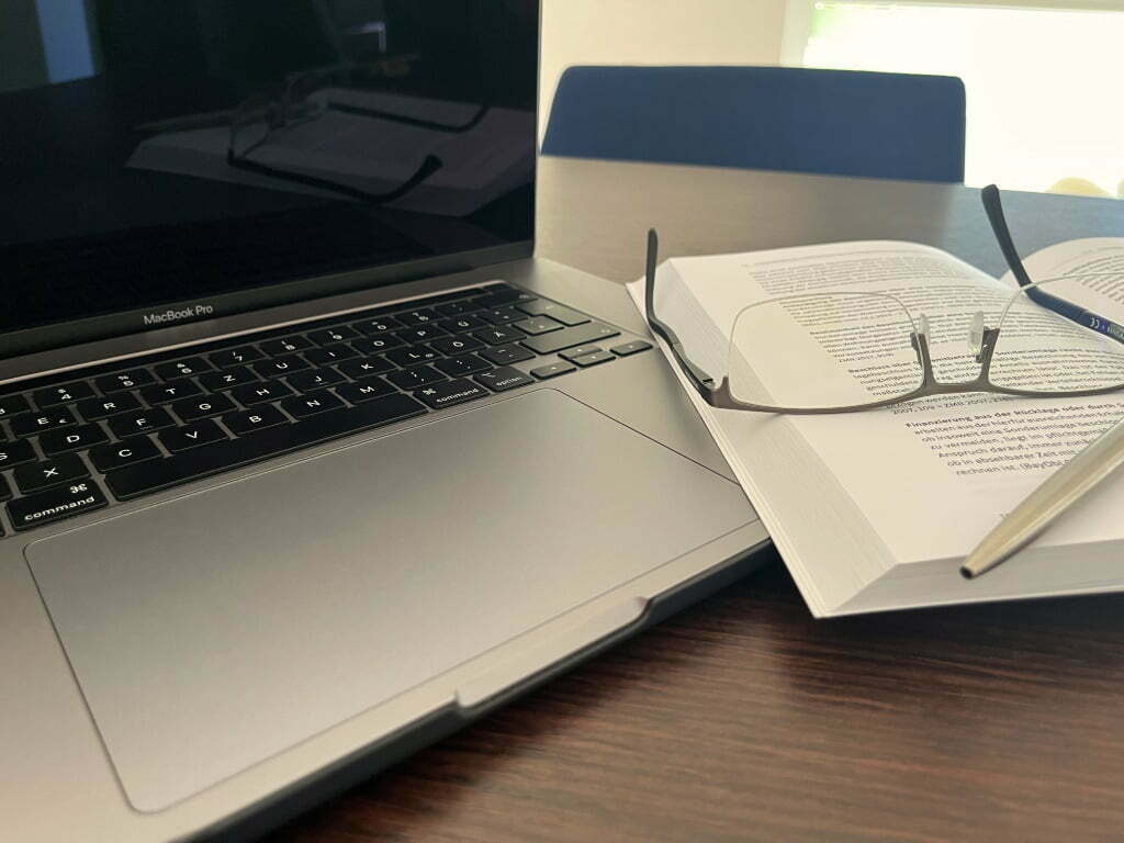 Laptop neben einem Buch auf dem eine Lesebrille und ein Kugelschreiber liegen